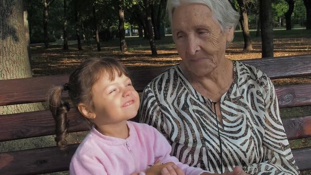 An elderly woman with a little girl outdoors. Grandmother with a little girl in a little nest sitting on a bench in the park.