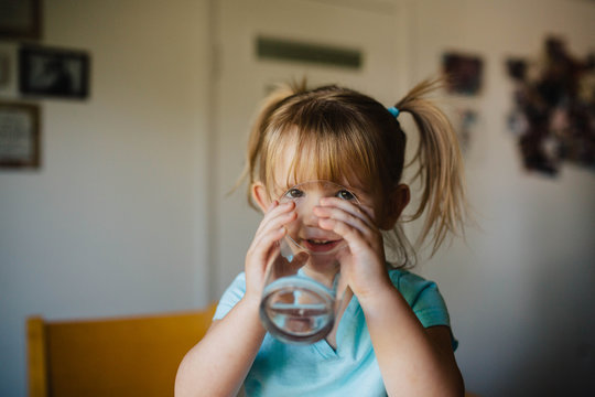 Toddler girl with pigtails sitting on the kitchen table drinking a big glass of water.