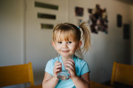 Toddler girl with pigtails sitting on the kitchen table drinking a big glass of water.