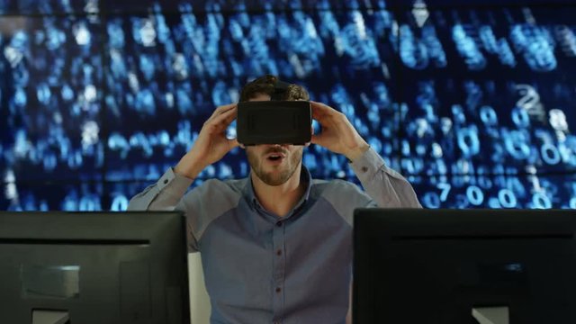  Engineer or computer programmer wearing virtual reality viewer at his desk with animated graphic of digital code in background