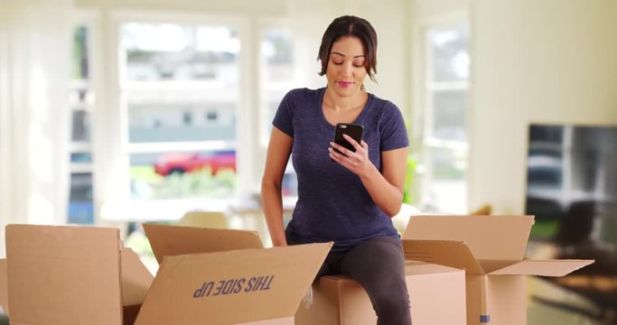 Frustrated Latina woman sitting on moving boxes inside living room, messaging someone on cell phone. Tired young female taking a break from packing up house. 4k 