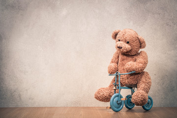 Teddy Bear sitting on old retro toy bicycle in front concrete textured wall background. Vintage...