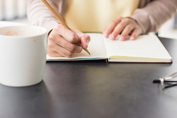 People hand writing with pencil on notebook near by coffee mug and glasses