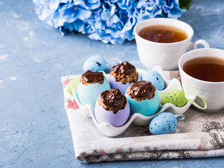 Obraz na płótnie Canvas Easter sweet cup cakes treets in colorful pastel egg shells with chocolate and hydrangea flowers on blue background. Festive holiday home made food with cups of tea