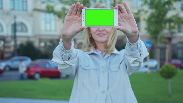 Young Woman staying in city park holds SmartPhone with pre-keyed green screen. Perfect for screen compositing. Made from 14bit RAW. 10bit ProRes 444