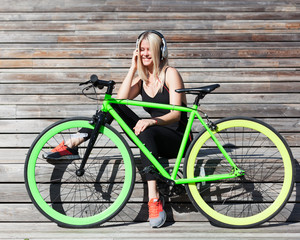 Obraz na płótnie Canvas Fitness princess with long blond hair girl in black sexy outfit and sneakers resting on wooden boards with fashionable fix bike listening to music on headphones on a sunny afternoon