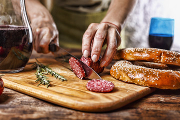 Traditional Italian red wine, salami, rosemary, bread. Close up of a person's hand cut salami on a kitchen board. - obrazy, fototapety, plakaty