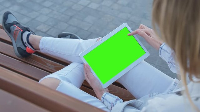 Young Woman in white jeans sitting on bench uses Tablet PC with pre-keyed green screen. Few types of gestures - scrolling up and down, tapping, zoom in and out. Perfect for screen compositing