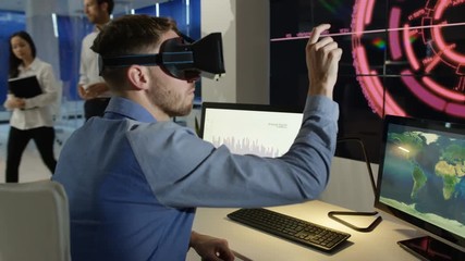Businessman interacting with a virtual reality headset in futuristic office, computer screens showing financial information & animated graphics - Powered by Adobe