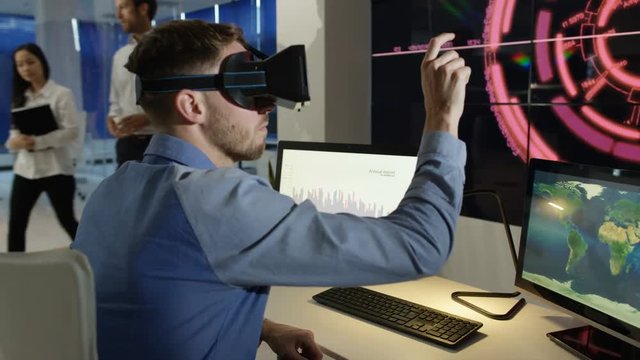  Businessman interacting with a virtual reality headset in futuristic office, computer screens showing financial information & animated graphics