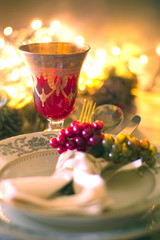 Christmas table setting, layout with cutlery, grapes decoration and a vintage red crystal goblet. Blurred celebration lights shining on the background for a bokeh effect.