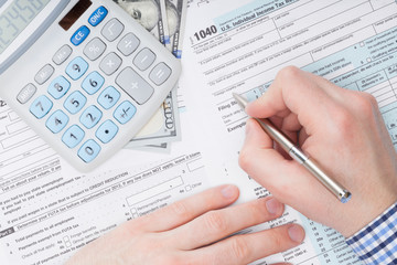 Close up shot of a male filling out US 1040 Tax Form next to calculator and 100 dollars banknote under it