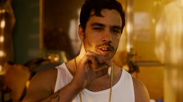 Smoking Brutal Gang Member with Gold Chain Looks into Camera with Defiance. Underground Drug Laboratory is in Background.