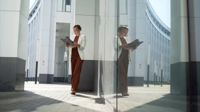 young business women in white suit standing and watching paper documents in black folder and go on camera. Modern white and glass building on background, wide angle shot