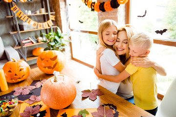 Lovely family cuddling, mum with two small blonde cheerful kids in decorated nice loft light room at home, desktop with cutted handmade pumpkins, colorful candies, fall leaves, garlands on window