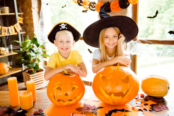 Fototapeta premium Close up cropped photo portrait of two small charming kids at halloween party indoors - blond small fairy and pirate, bonding, on top of hand cutted pumpkins, cheerful toothy smiles, indoors at home
