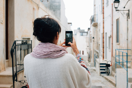 Brunette Woman in White Poncho Taking Cellphone Photo in Whitewashed Italian Village