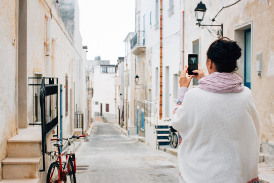 Southern Italy - Brunette in White Poncho Standing in Quaint Whitewashed Village and Taking Smartphone Photo