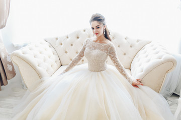 Wedding. Bride in beautiful dress sitting on sofa indoors in white studio interior like at home....