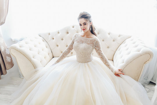 Wedding. Bride in beautiful dress sitting on sofa indoors in white studio interior like at home. Trendy wedding style. Young attractive caucasian brunette model like a bride