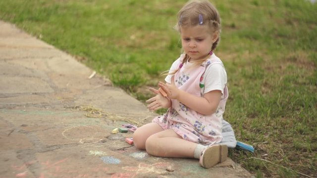 Portrait of a cute little girl dressed in pink overalls, the child draws colorful chalk on the path next to the green grass on a summer sunny day.