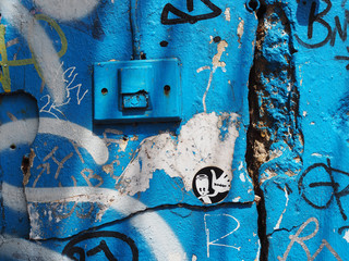 Blue painted old peeled cracked grainy grungy texture concrete wall with bell ring alarm switch panel, and messy graffiti spraying