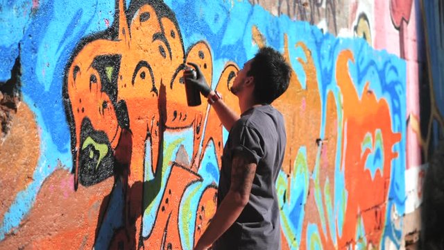Handsome Talented Young Boy making a colorful graffiti with aerosol spray on urban street wall. Cinematic toned slow motion footage. Creative art. Gimbal shot. Side view