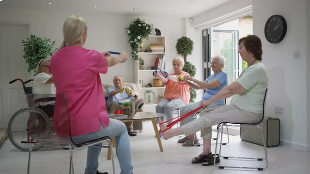 Young Nurse Showing Elderly Group Some Exercises With A Resistance Band In Nursing Home.