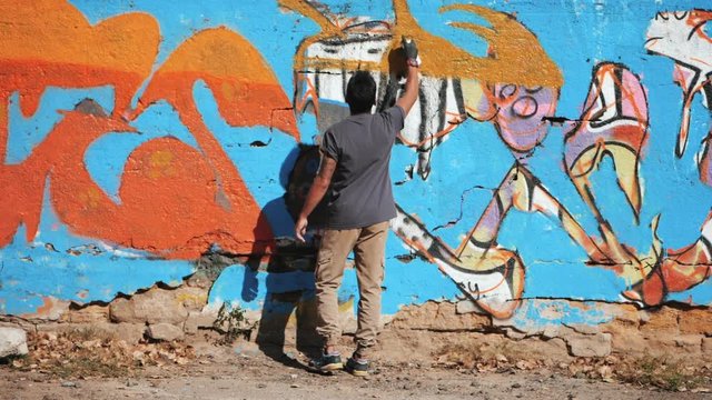 Handsome Talented Young Boy making a colorful graffiti with aerosol spray on urban street wall. Cinematic toned slow motion footage. Creative art. Back view, overall plan