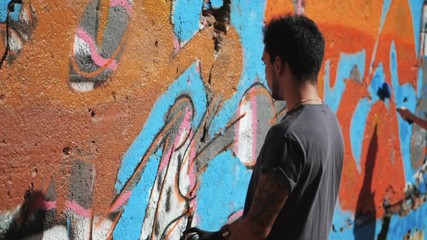 Graffiti Artist Painting On The Street Wall. Handsome Man with aerosol spray bottle spraying with colorful paint, Urban Outdoors Art Concept. Slow motion.
