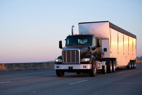 Dark big rig semi truck with bulk trailer running on highway in sunset light