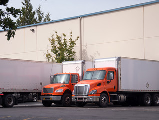 Orange middle semi trucks with box trailers stand in warehouse dock under unloading