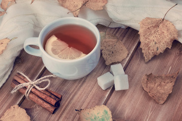 Tea with lemon in white cup, cinnamon sticks, sugar and autumn yellow leaves on wooden background