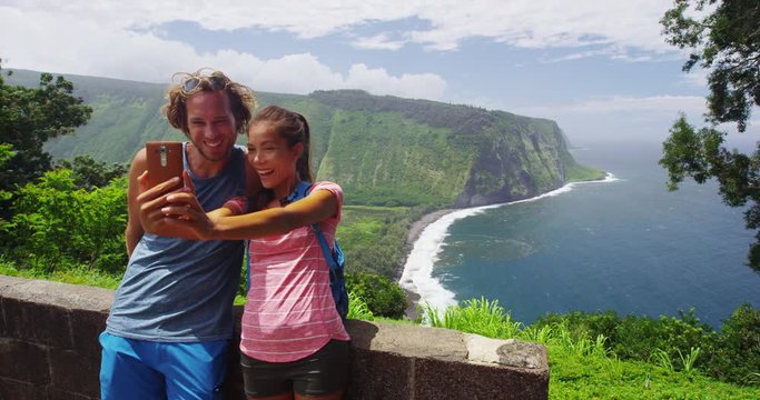 Tourists couple taking phone selfie photo at Waipio Valley Lookout Big Island Hawaii landscape. Couple on travel by Hawaiian nature at famous tourist destination on Big Island, Hawaii, USA. RED EPIC.