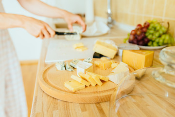 Close up photo cheese plate - woman's hands cutting different kinds of cheeses in counter at kitchen home interior.