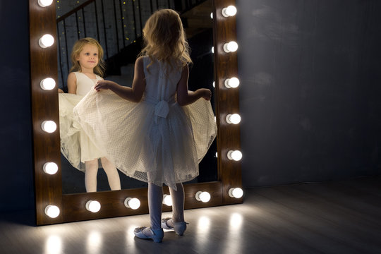 A girl, a child, a model in a beautiful dress is standing by the mirror and considering her reflection. Portrait, child, mirror.