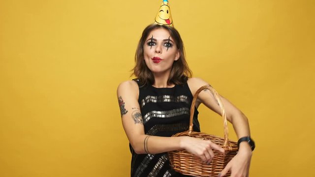 Tricky lady with halloween make up and holiday cap eating candies in secret and laughing isolated over yellow