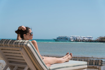 Girl sunbathing on a lounger on the beach. In the background a pier and yacht are visible