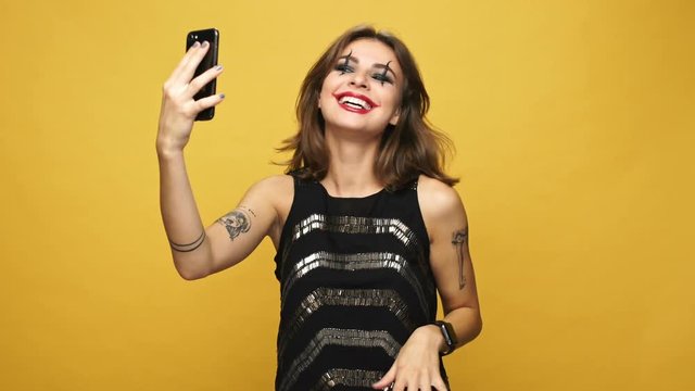 Beautiful happy woman with creepy halloween make up making selfie with scary funny emotions on smartphone isolated over yellow