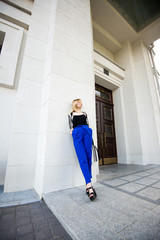 Стильная, модная девушка со светлыми волосами стоит возле белого здания в синих брюках и черной майке 