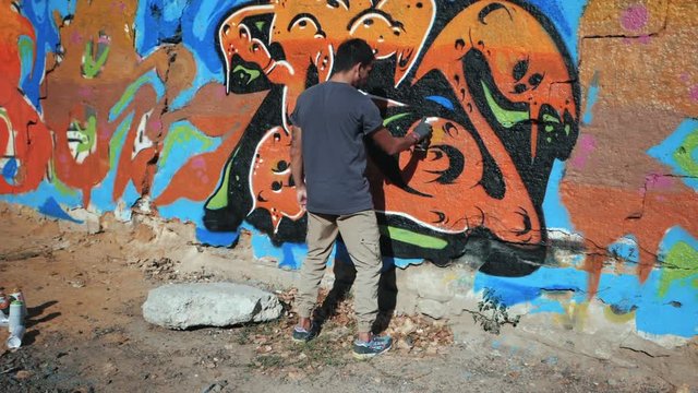 Graffiti Artist Painting On The Street Wall. Handsome Man with aerosol spray bottle spraying with colorful paint, Urban Outdoors Art Concept. Slow motion. Gimbal shot