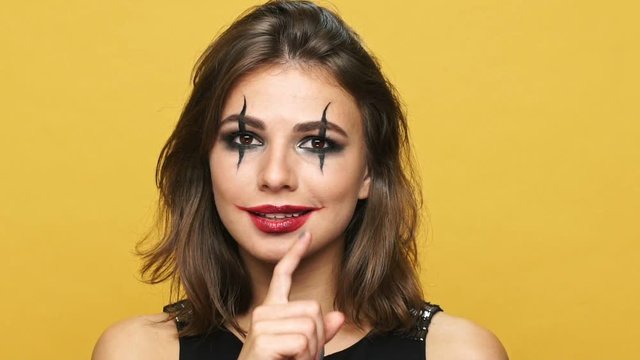 Cheerful creepy woman with dark make up showing ok gesture and winking isolated over yellow