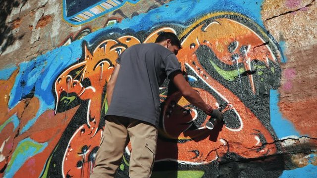 Graffiti Artist Painting With Aerosol. Man with spray bottle. Slow Motion. Young urban painter drawing colorful graffiti on the urban street wall at summer sunny day. Back view, overall plan