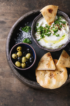 labneh middle eastern lebanese cream cheese dip with olive oil, salt, herbs served with olives, traditional pita bread on terracotta plate over dark texture wooden background. Top view with space