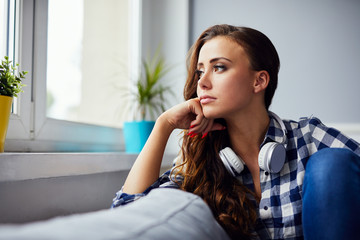 Fototapeta premium Sad melacholic young woman sitting on sofa at home and looking through the window