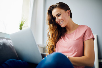 Portrait of a happy beautiful young woman smiling and using laptop while sitting on sofa in living room