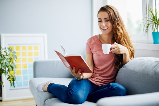 Cheerful attractive young woman reading a book and drinking coffee, sitting on sofa in home