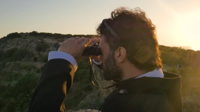 Man in elegant suit with briefcase looking through binoculars on top of a cliff on an island