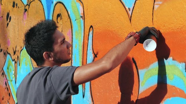 Street artist painting colorful graffiti on generic wall - Modern art concept with urban guy performing and preparing live murales with aerosol color spray - Sunny afternoon. Slow motion. Side view