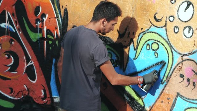 Graffiti Artist Painting With Aerosol. Man with spray bottle. Slow Motion. Young urban painter drawing colorful graffiti on the urban street wall at summer sunny day.